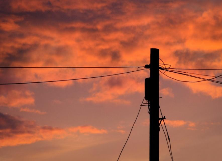 The dark shadowed outline of a utility post with power lines extending off to the side and towards the ground in front of an orange-gold, cloud-filled sky right before sunset.