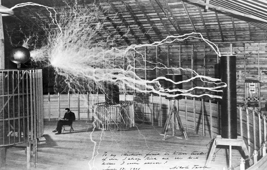A black and white photograph of the inside of Wardenclyffe Tower, a station Tesla built to transmit messages across the Atlantic. He sits in the background, to the left, under the tall, wooden rafters. In the foreground are two machines transmitting electricity to one another, in visible, lightning-like, electrical currents.
