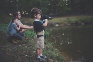 A father crouches beside a pond, his son, crouching beside him, rests his head against the father’s shoulder, while a few steps away a younger son stands with a fishing rod in his hand, watching the water carefully.