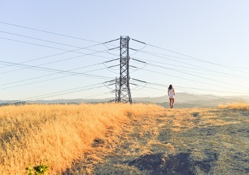 On a sunny day, in a landscape with yellow grass on a hill there is a person with hands in their pockets walking away from the camera, towering above the person is an electric tower with cables and transformers.