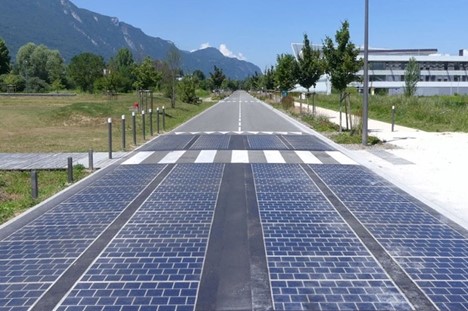 a black reflective solar panel roadway, stretching into the distance toward a mountain and a blue sky; there are trees and grass on one side of the road in a small building with a line of trees on the other.