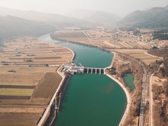 Aerial view over a hydroelectric power station on a river surrounded by fields, roads, and mountains.
