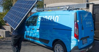 A baby blue Sunrun-branded van parked tightly against a garage in the driveway of a residential house, as a Sunrun employee carries a solar panel carefully against their shoulder.