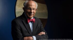 Dr. Warren M. Washington smiling for a portrait, wearing glasses, in a suit and a colorful red bowtie, standing in front of a blue wall and wall art.
