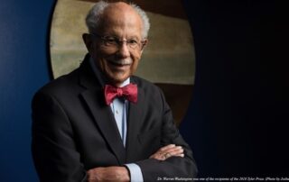 Dr. Warren M. Washington smiling for a portrait, wearing glasses, in a suit and a colorful red bowtie, standing in front of a blue wall and wall art.