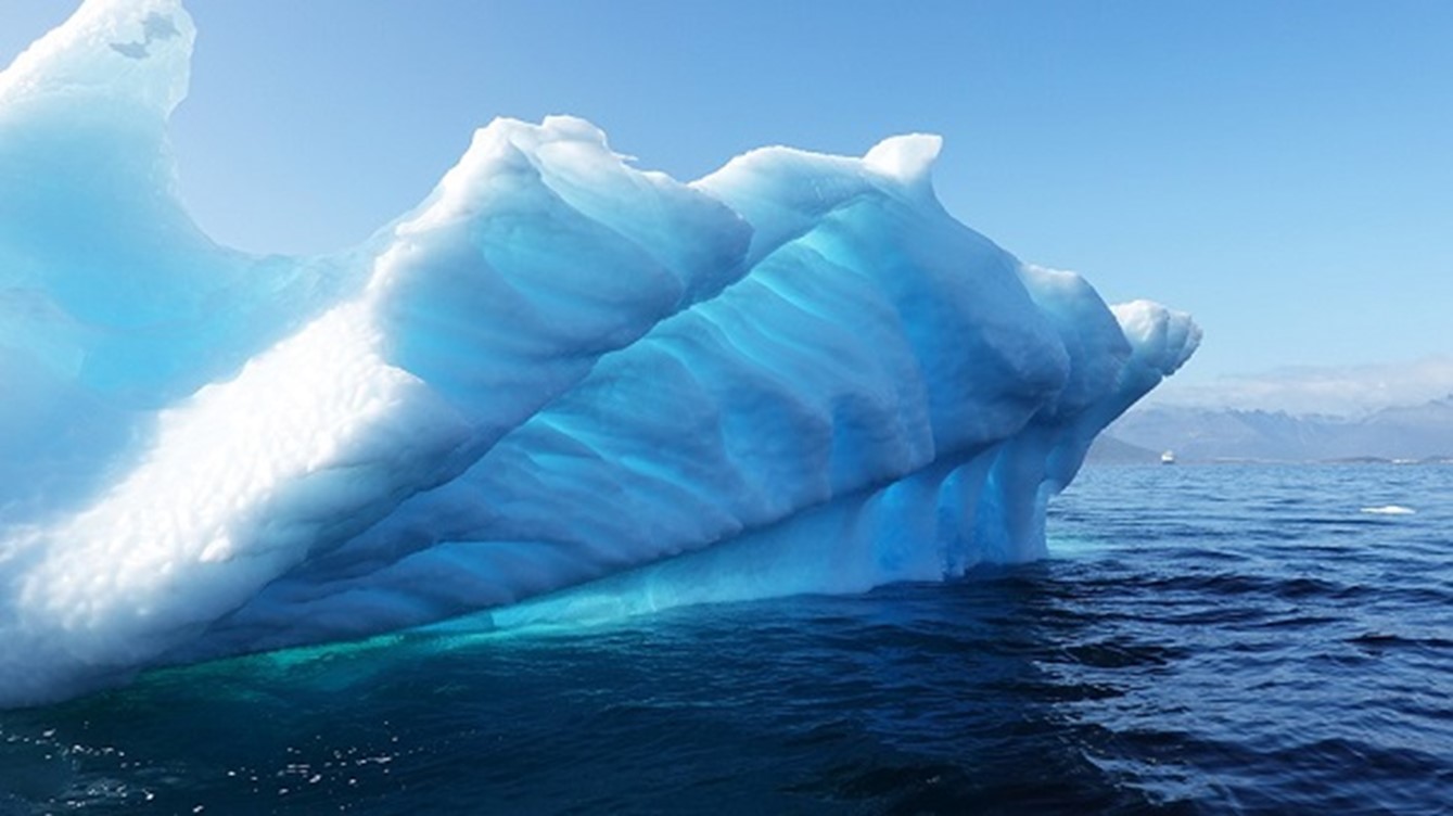 A photo of a large iceberg floating in the ocean with sun shining on the ice.