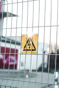 A square yellow sign attached to a metal fence with a high voltage symbol as warning.