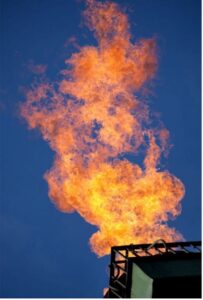 A photo of fire burning so intensely on top of a steel ledge. The shot was taken from the ground up and against a clear and blue sky.