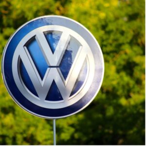 A blue and silver sign with the Volkswagen logo on it. The background is a forest of trees blurred out.