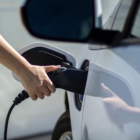 a hand holds a black nozzle to the side of a white electric vehicle to charge it.