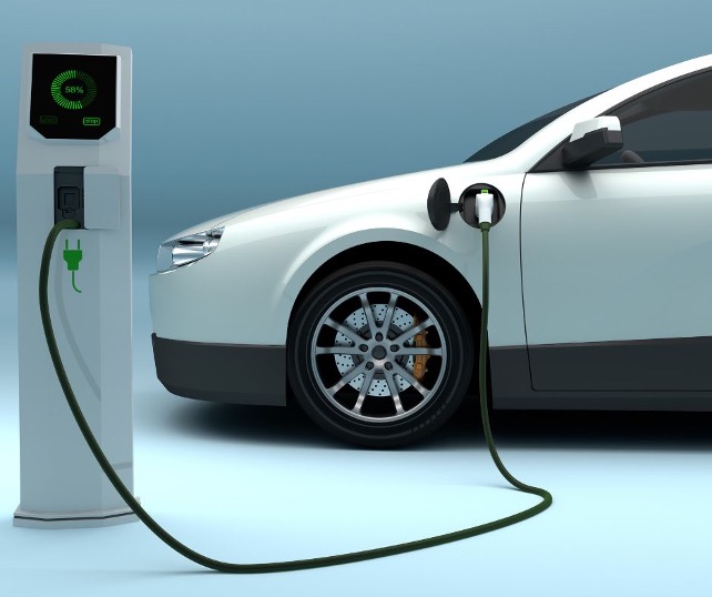Photo of a white and black electric vehicle parked next to a futuristic-looking white charging station and plugged into it by a cord. The background is just a plain white room.