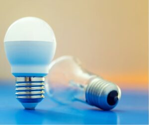 Photo of two light bulbs, one that is standing is an LED and the other that is lying down is incandescent. The background is a blurred gradient image with orange and blue.