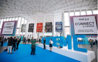 Pictured is a large indoor area with a high ceiling, big windows, and an attractive modern design with white architectural shapes. Big blue letters at the center of the atrium spell “CWIEME.” Lots of people are talking and walking around the space. Banners advertising different companies are hanging from the ceiling.